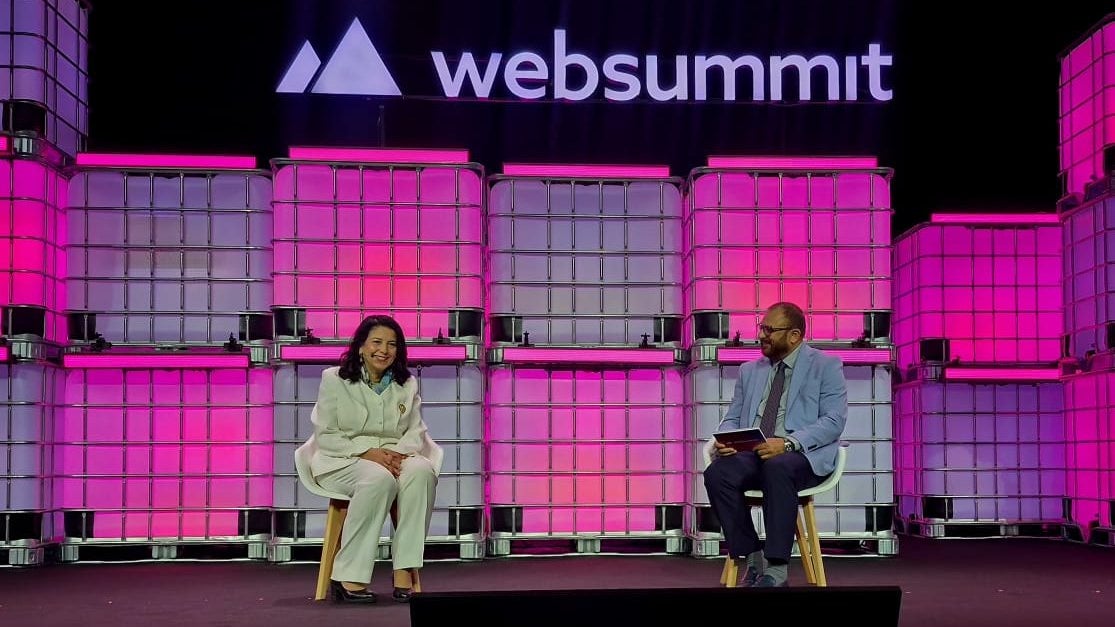 A woman in a white suit sits on stage next to a man in a suit, both speaking at the Web Summit event with pink-lit containers behind them.