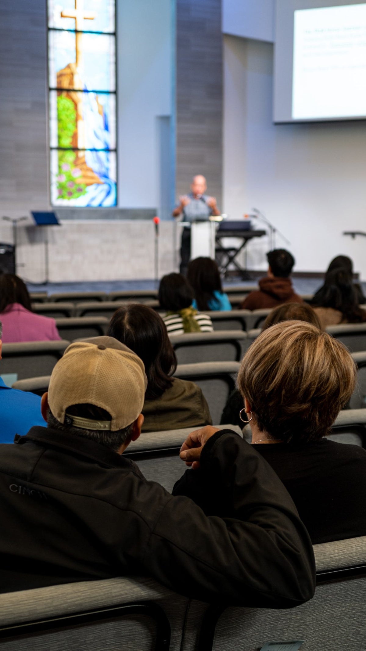 A photography of two people seated in a church service as a man speaks on stage.