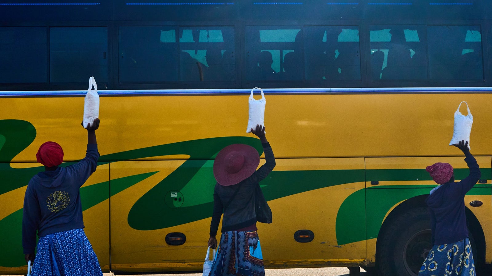 Three women holding bags up to the window of a large yellow bus, with two wearing patterned skirts and the third in a hat, set against a clear sky.