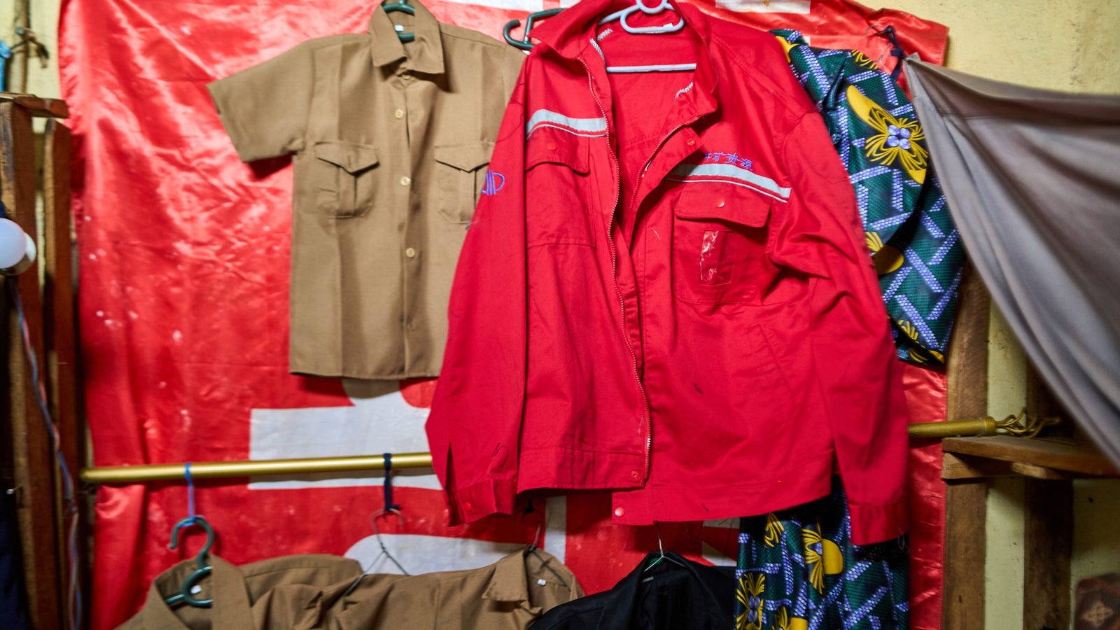 A red jacket and a tan short-sleeved shirt are hanging on hangers against a backdrop of a red and white cloth; additional colorful patterned cloth is visible in the background.