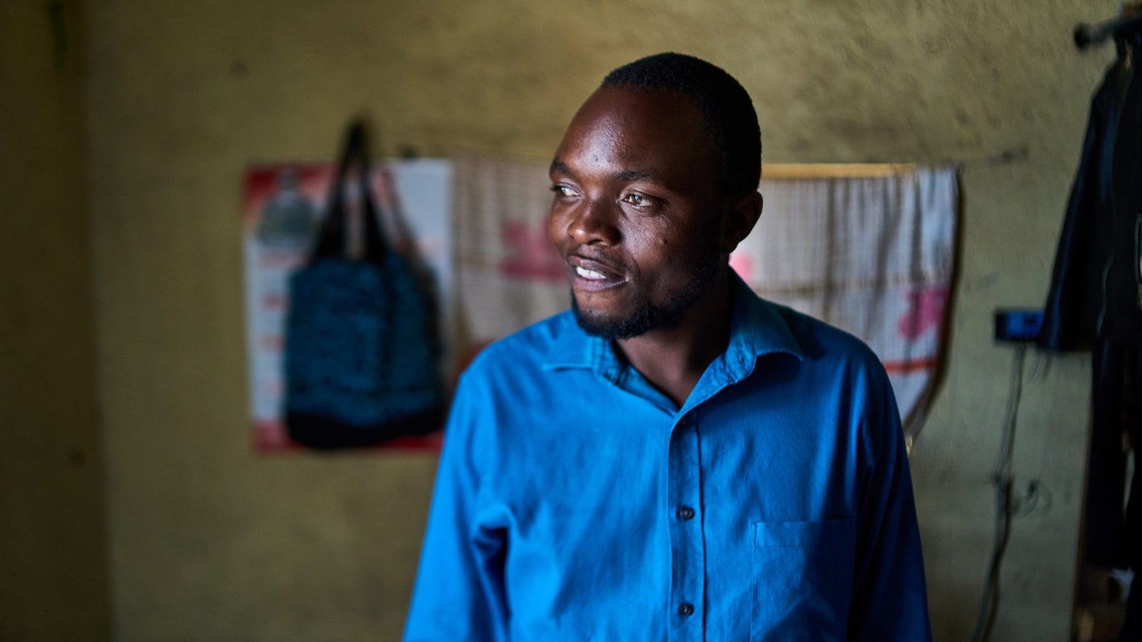 A man wearing a blue shirt stands in a dimly lit room with a textured wall, smiling softly as he looks to the side. In the background, there is a patterned bag hanging, along with a decorative cloth and electrical wires.