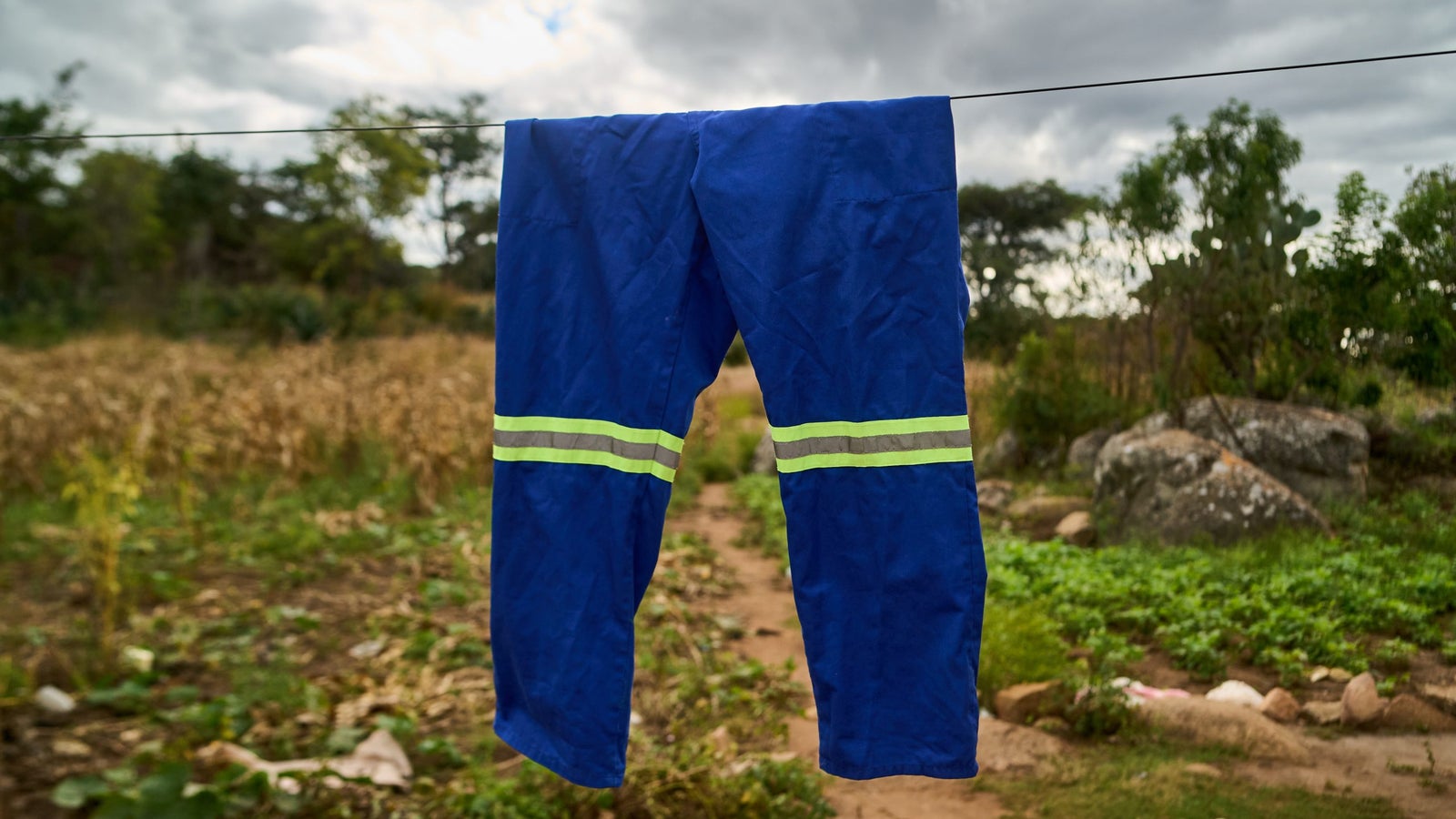 A pair of blue pants hanging on a clothesline, featuring reflective stripes in yellow and gray, with a rural landscape of crops and rocks in the background under a cloudy sky.