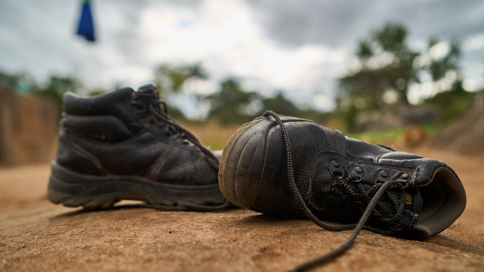 A close-up view of two black boots resting on a sandy surface, with one boot slightly turned towards the camera, showcasing its worn laces and rugged design; the background features a blurred landscape with patches of greenery and cloudy skies.