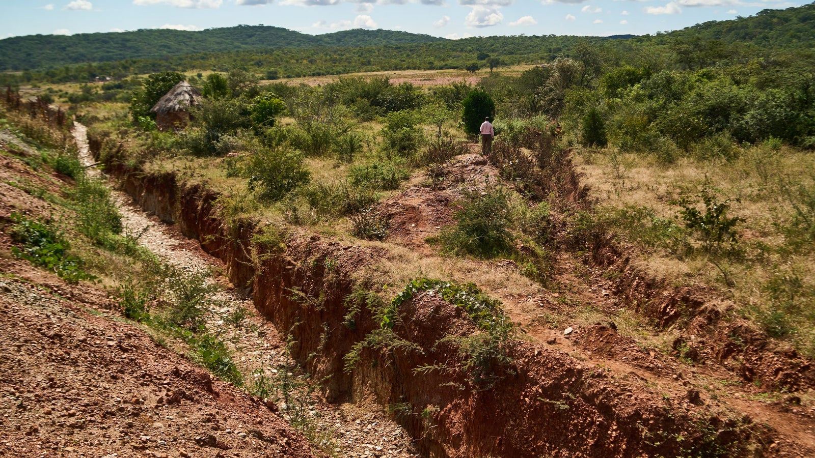 A person walking along a dry, rocky trench in a rural landscape dotted with shrubs and a thatched hut, set against a backdrop of rolling green hills and a partly cloudy sky.