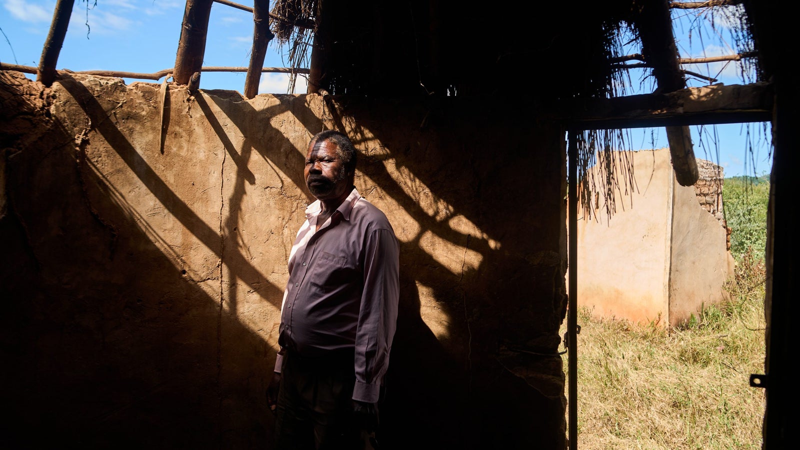 A man stands against a textured earthen wall inside a rustic structure, illuminated by sunlight filtering through the roof, casting dramatic shadows. In the background, there is a view of a grassy area and another building visible through an open doorway.