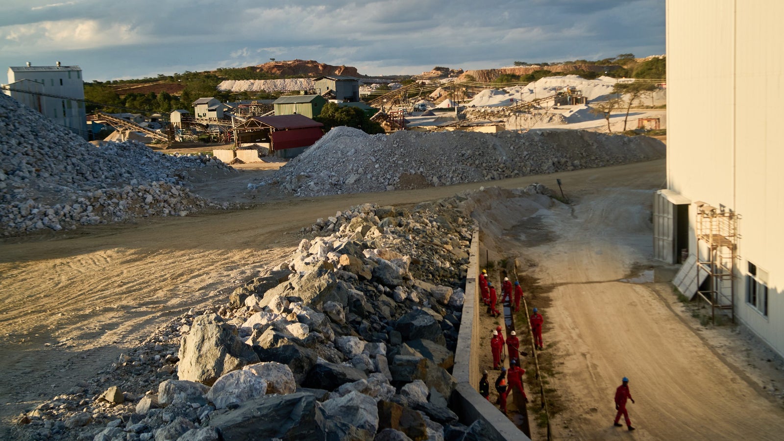 A mining site with large piles of rocks and gravel, featuring several workers in red uniforms walking along a dirt pathway, and various industrial buildings in the background under a partly cloudy sky.