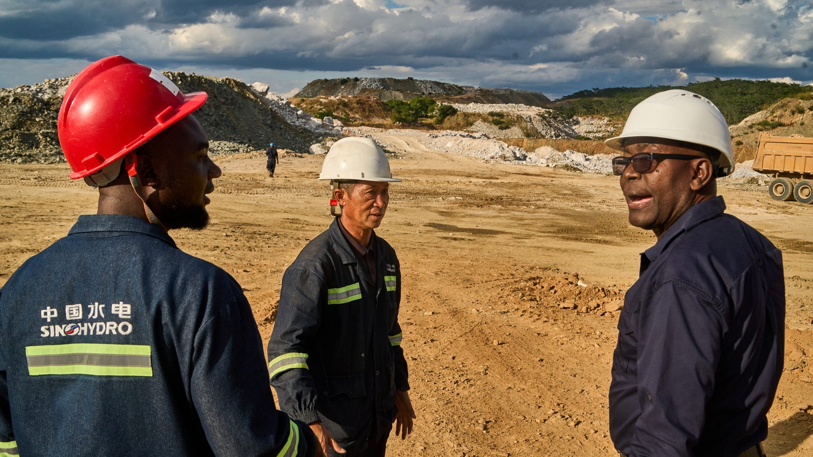 Three construction workers in safety helmets and uniforms converse on a dirt site with machinery and hills in the background, under a partly cloudy sky.