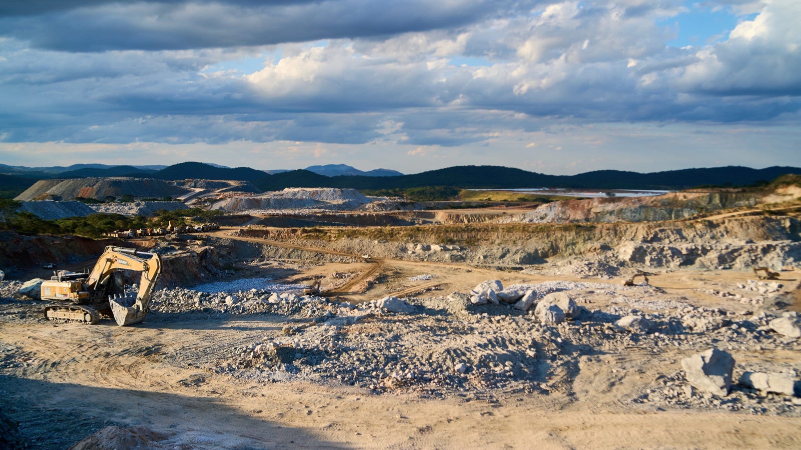 A view of an open-pit mining site with heavy machinery, including an excavator, amidst a landscape of rock piles and gravel, under a partly cloudy sky.