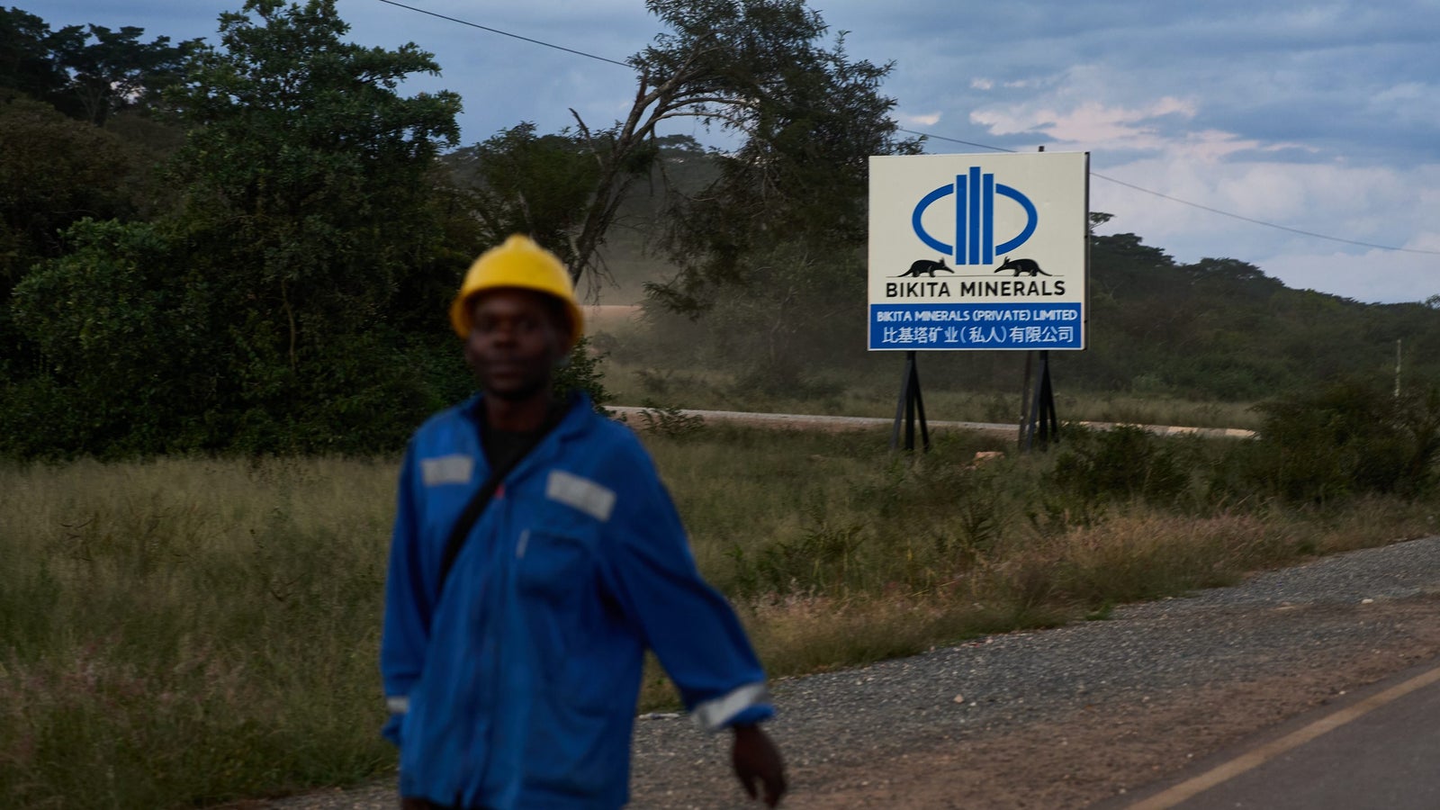 A man in a blue work uniform and yellow hard hat walks along a gravel road near a sign for Bikita Minerals, which displays the company name in English and Chinese, surrounded by greenery and a cloudy sky.