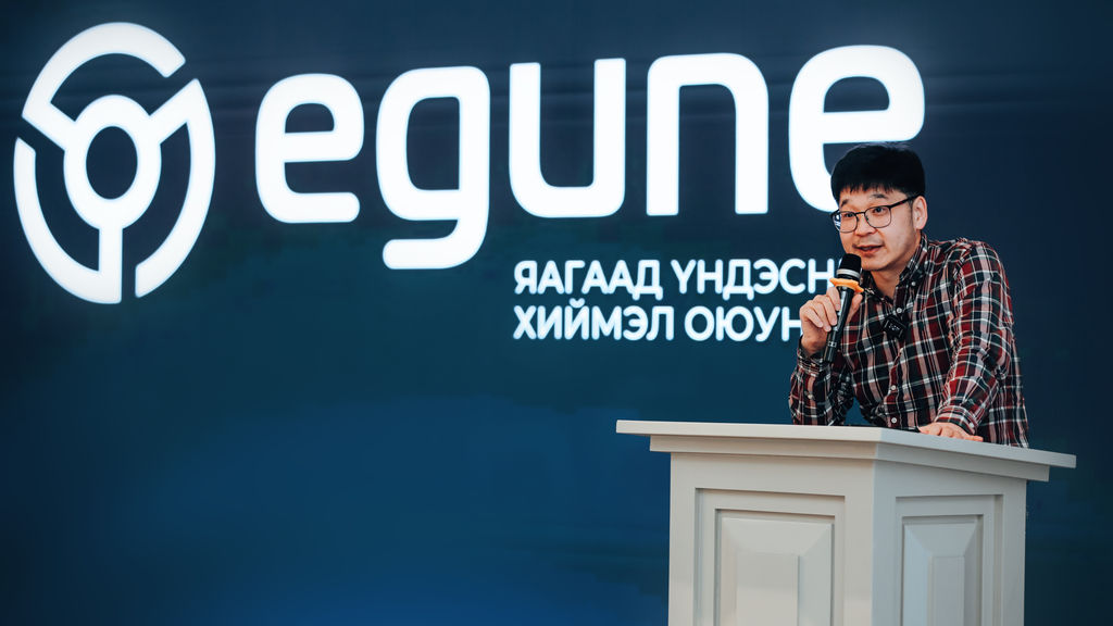 A man in a checkered shirt speaks into a microphone at a podium with a logo of 'egune' displayed in the background, on a blue backdrop featuring text in Mongolian below the logo.