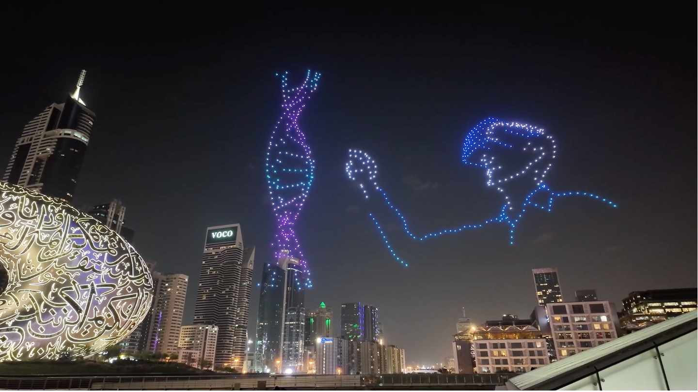 A nighttime view of a city skyline featuring a captivating light display in the sky, depicting a large outline of a person holding a microphone next to a stylized DNA helix, illuminated in vibrant colors against a dark backdrop.
